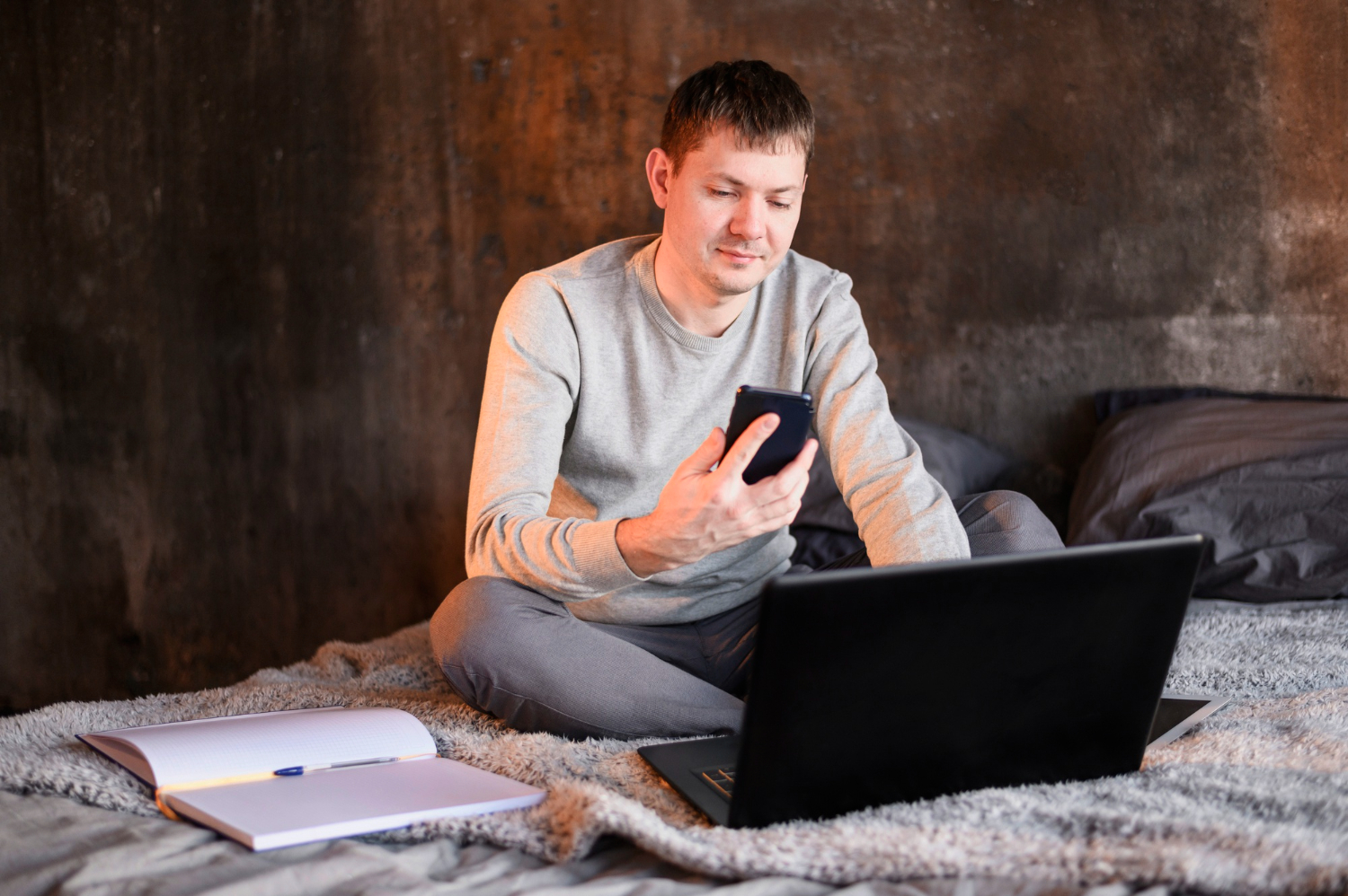 A man sits on a bed with a laptop and a phone